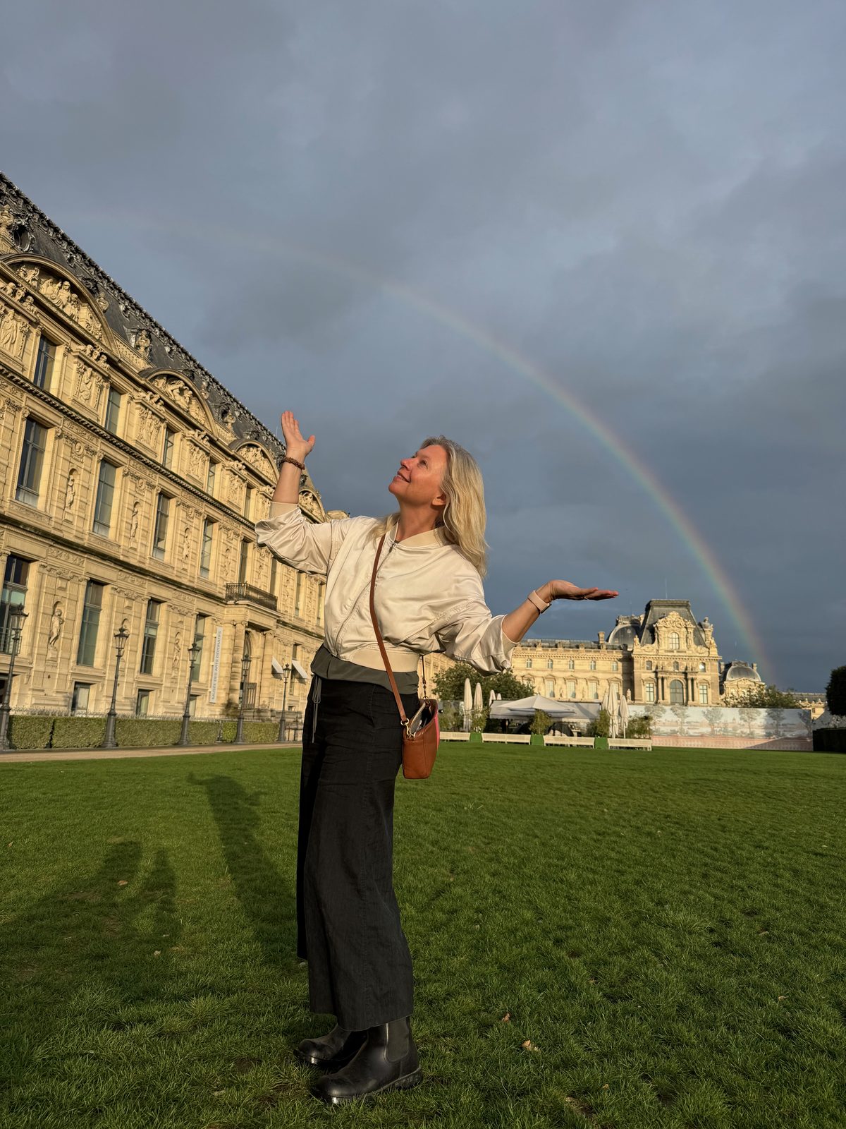 Maggie Langrick outside the Louvre in Paris, arms open wide, with a rainbow arcing across a dramatic stormy sky