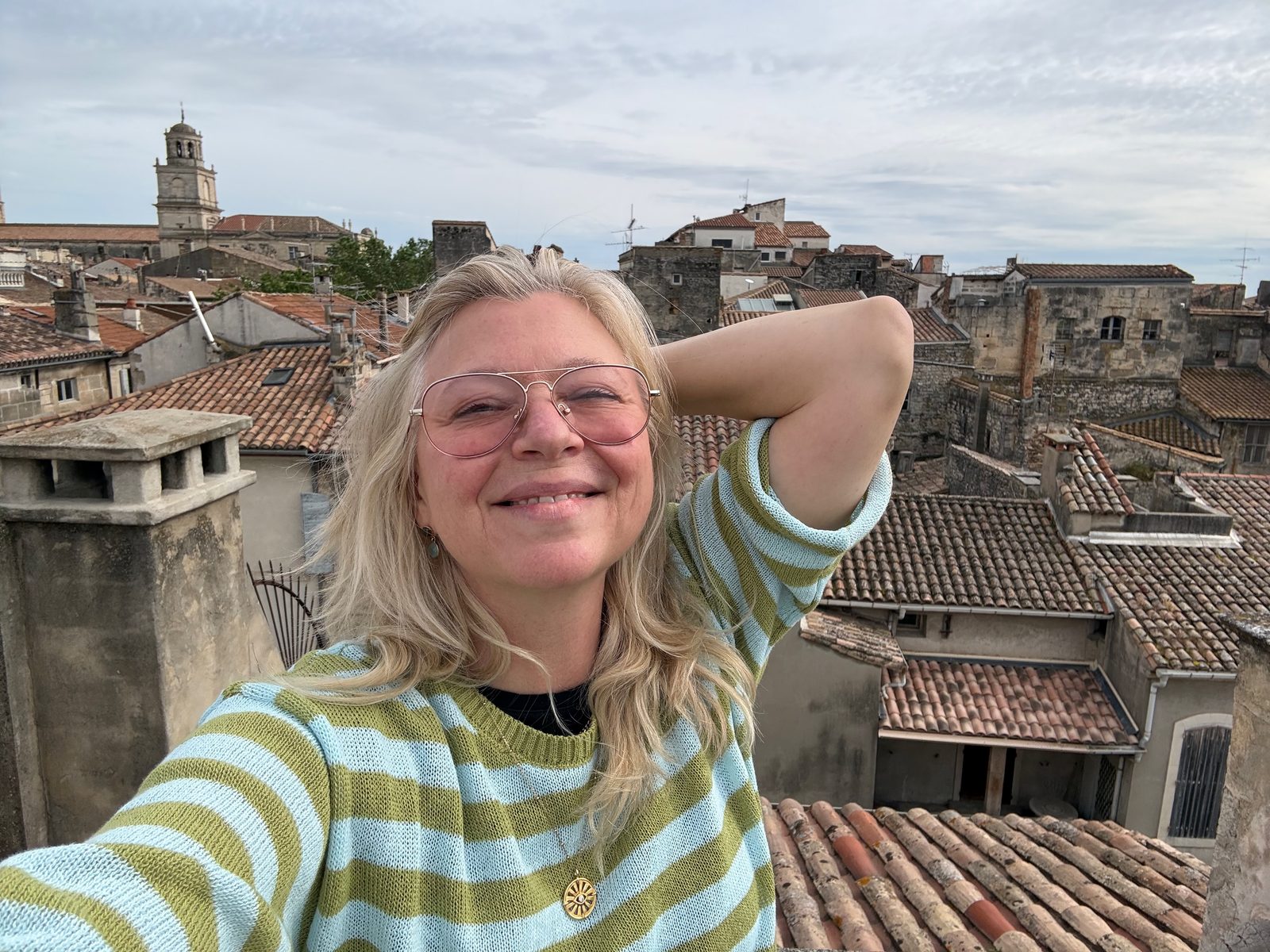 Maggie Langrick on a rooftop in Arles, France, smiling against a backdrop of terracotta rooftops and a church tower